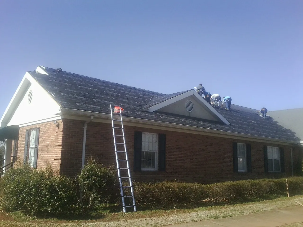 Crew tearing shingles off a brick ranch roof with a ladder set — Emergency Roof Repair work by our crew in Mcintosh