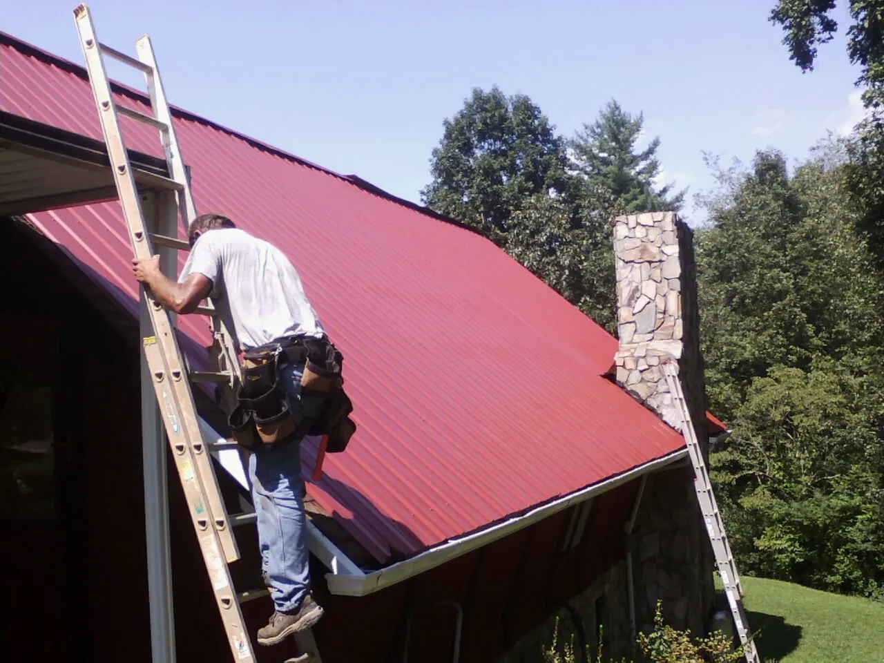 Roofer on a ladder beside a red metal roof and stone chimney — Soffit & Fascia Repair by Roofing Brothers in Mcintosh